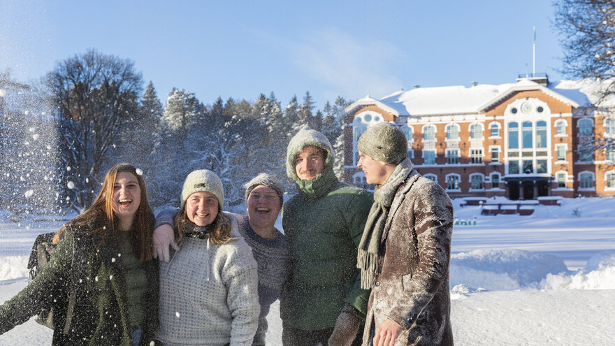 Fem studenter smiler foran Urbygningen. Det er mye snø ute.