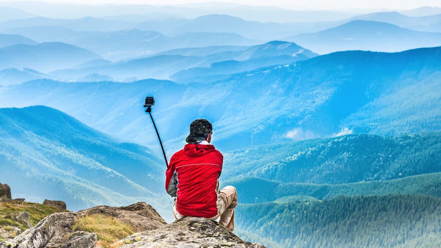 Person på fjelltopp, ser på utsikt og holder i selfie-stang